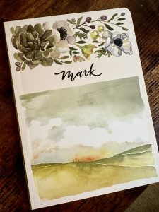 Scripture journal on wooden table with watercolor artwork on cover featuring a sunset behind three crosses on bottom of cover, florals on the top of the cover, and "mark" written in cursive on the middle of the cover. 